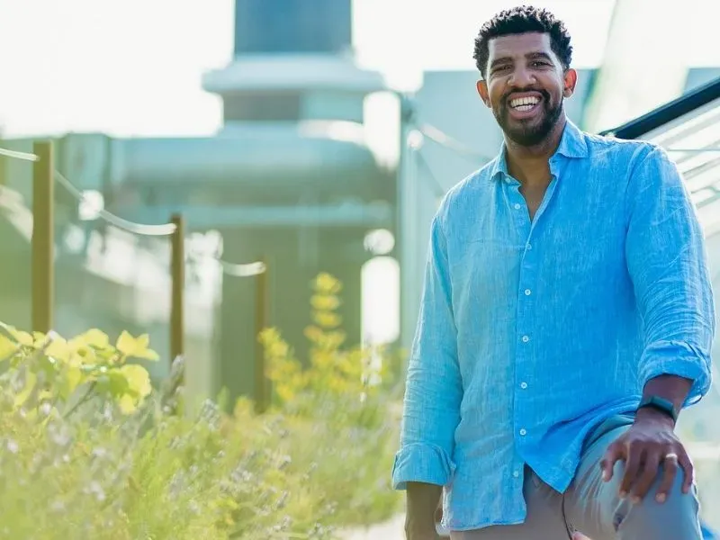 Smiling man in a blue shirt standing relaxed outside with greenery and modern architecture in the background